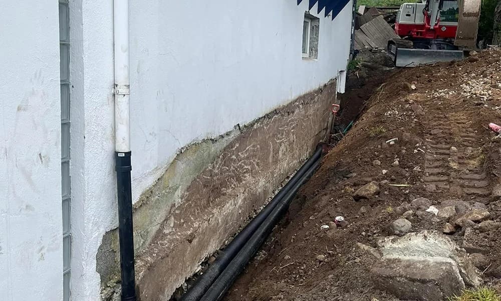Avdekket grunnmur langs en grøft med rør, jord og gravemaskin i bakgrunnen.