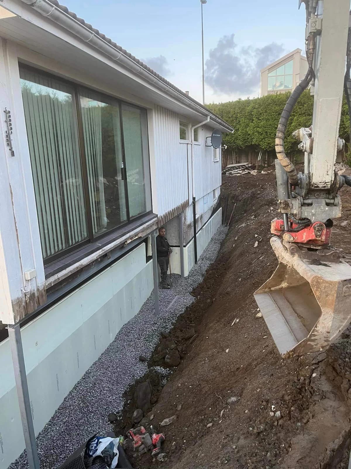 Gravemaskin og person ved en lang åpen grøft, isolert grunnmur og pukk.
